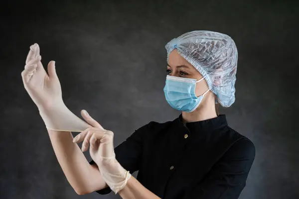 Caucasian young woman doctor or nurse in black uniform and protective mask putting protective glove isolatedon dark. Healthcare concept