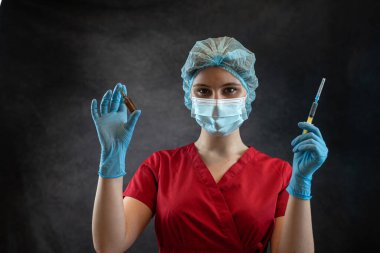 Doktor kadın kırmızı üniformalı eldivenler giyiyor. Şapka maskesinde elinde siyah bir şırınga var. Stüdyo portresi. Sağlık çalışanı. 