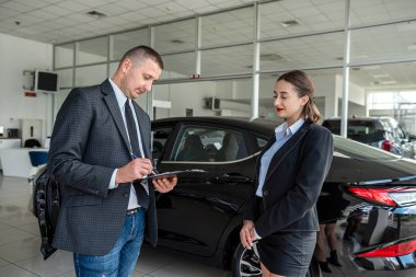 young manager in suit holding clipboard with female client  talking to sales modern car at showroom