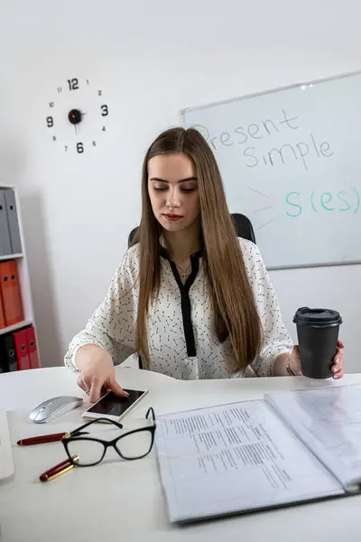 Smiling woman teacher  sitting at work desk with laptop and explaining grammar rules near whiteboard