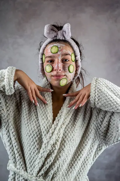 Beautiful young woman receiving skin care procedure with white cream and cucumber. Concept of selfcare and healthy lifestyle