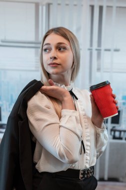 Takım elbiseli iş kadını elinde kahve tutuyor ve iş merkezinin koridorunda tek başına dinleniyor. Yemek molası.