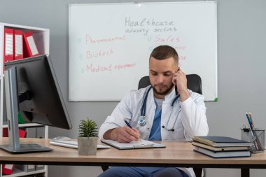 Beyaz üniformalı, steteskop işi olan beyaz bir doktor, çalışma masasında oturuyor.