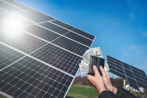 hand holding dollar bills that look out of a wallet on a background of solar panels. saves money. using solar energy. green electricity