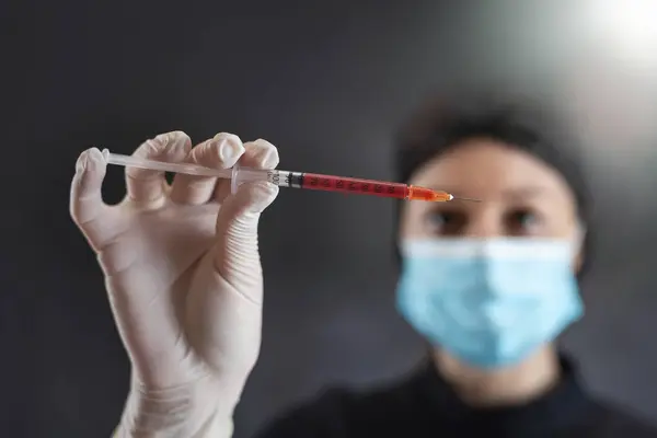woman doctor in black uniform mask and gloves holding blood sample  or red liquid in syringe isolated on dark