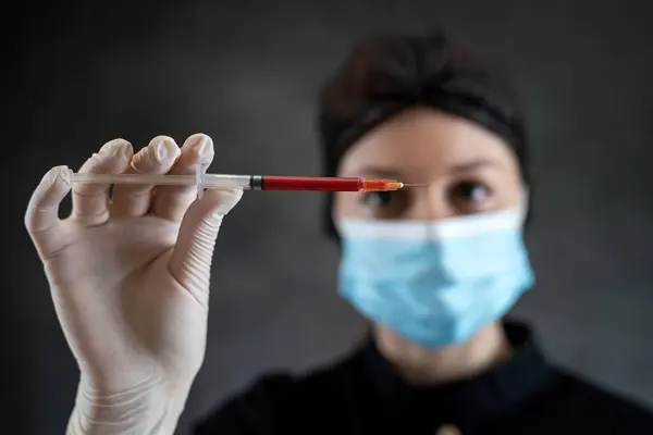 woman doctor in black uniform mask and gloves holding blood sample  or red liquid in syringe isolated on dark