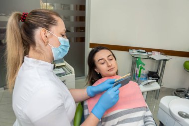 Diş hekimi, beyazlatma tedavisi için diş örneklerini tutuyor ve genç bayan hastasına bir gölge seçiyor. Diş sağlığı ve sağlık hizmetleri kavramı 