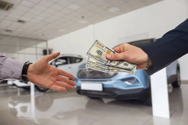 man gives dollar cash to dealer man when buying a car at modern showroom. Deal of buy or rent new auto