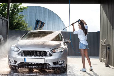 Zayıf bir kadın arabasının köpüğünü yüksek basınçlı su tabancasıyla yıkar. Araba yıkama servisi