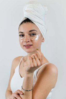 Havlu giymiş genç beyaz bir kadın beyaza izole edilmiş yüzüne hidrojel bantlar uyguluyor. Cilt bakımı kozmetik prosedürleri