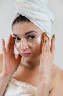 Beyaz havlu giyen genç bir kız gözlerinin altında kozmetik bantlar kullanıyor. Beyazlar içinde izole edilmiş. Taze yüz derisi, bakım