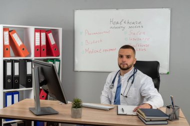 Beyaz üniformalı, steteskop işi olan beyaz bir doktor, çalışma masasında oturuyor.