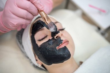 Güzellik uzmanı, kaplıca salonundaki genç ve güzel kadınlara siyah maske takıyor. Yaşlanma karşıtı kozmetik prosedür kavramı 