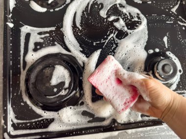 Kadın sünger bezini beyaz köpükle temizliyor ve deterjan da mutfak ocağını yıkıyor. Ev temizliği kavramı