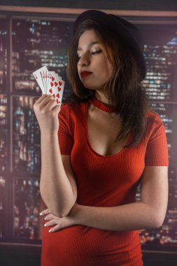 Poker masasının yanında elinde poker kartları olan kırmızı baştan çıkarıcı elbiseli güzel siyah saçlı kız. Poker oyunu kartları