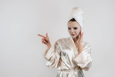 Beyaz havluyla genç bir kadın beyaz arka planda izole edilmiş nemlendirici beyaz bir maske kullanıyor. Yaşlanma önleyici prosedür. 