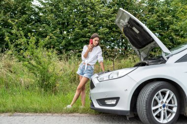 Endişeli kadın, yaz tatili sırasında bozuk bir arabanın kaputuna bakıyor. Servis otomatiği için yardıma ihtiyacın var