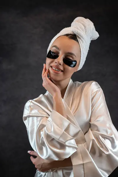 stock image Attractive woman wear towel with eye patches posign at camera isolated on black. Cosmetic procedure for reducing wrinkles skin under eyes