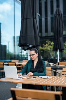 Güzel beyaz takım elbiseli iş kadını, şehir caddesindeki modern binanın dışındaki masada otururken dizüstü bilgisayar kullanıyor. 