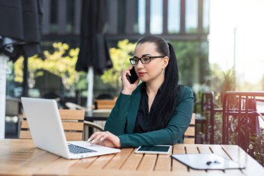  İş kadını laptopta çalışıyor ve açık kahve masasında telefonla konuşuyor.