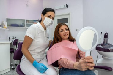 beautiful young female patient with a mirror in her hands examines beautiful treated teeth next to a female doctor. the concept of treatment in dentistry. dental health