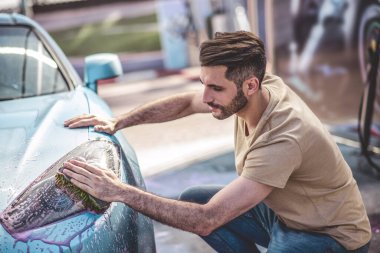 Ciddi bir genç araba tamircisi araba farlarını deterjan ve mikrofiber süngerle yıkıyor.