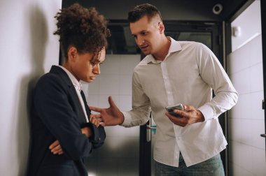 Açıklama. Beyaz, orta yaşlı, akıllı telefonlu bir adam ofiste kameraya karşı duran üzgün mulatta kadınına bakıyor.