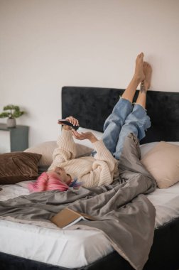 Pink-haired girl lying on her back on the bed with the smartphone in her hands