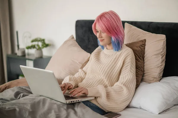 Focused young female seated in bed wrapped up in the blanket typing on her laptop