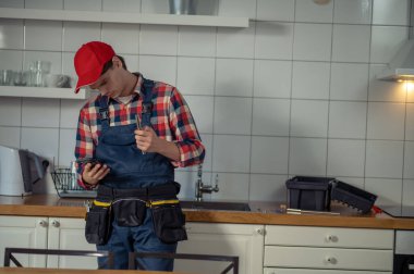 Serious focused repair technician standing at the kitchen counter and looking at the smartphone screen