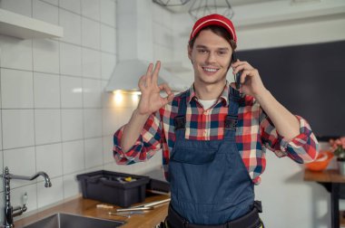 Waist-up portrait of a contented plumbing technician making the OK sign during the phone call in a customer kitchen
