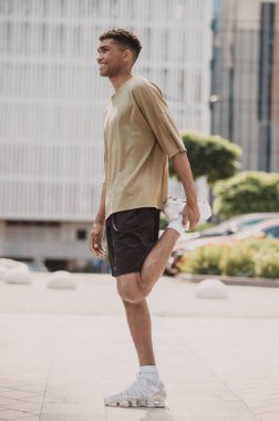 Side view of a smiling cheerful African American sportsman performing the standing quad stretch outdoors