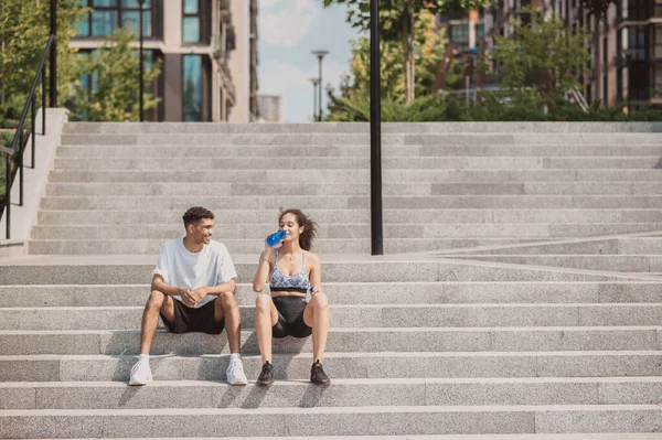 Young woman seated on the concrete steps and drinking water in the presence of her cheerful fitness partner