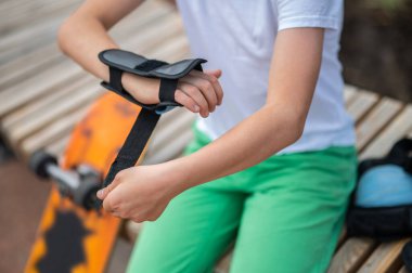 Cropped photo of a skateboarder seated on the wooden bench putting the wrist guard on the carpus