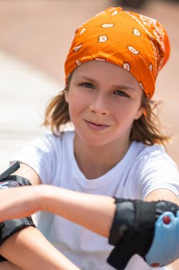 Serious cute fair-haired preadolescent boy with the elbow pads sitting outdoors on a sunny day