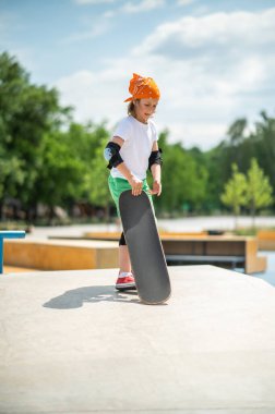 Full-size portrait of a young skateboarder in the elbow pads gripping his skateboard with one hand