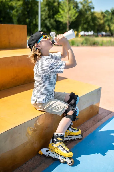 Roller skater in the sunglasses and knee pads sitting on the bench and drinking water