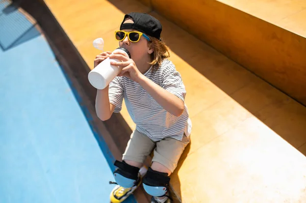 Boy in the knee pads and sunglasses sitting on the bench and drinking water from the plastic sports bottle