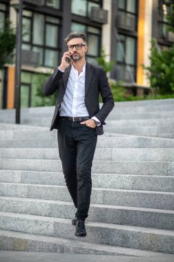 Telefon var. Takım elbiseli ve beyaz gömlekli bir adam sokakta yürüyor ve telefonda konuşuyor.