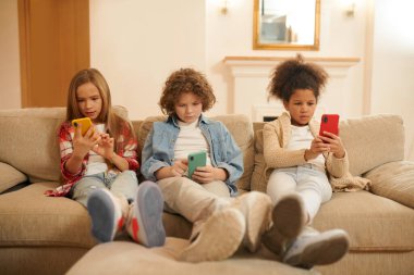 On internet. Kids sitting on the sofa with gadgets in hands and looking involved