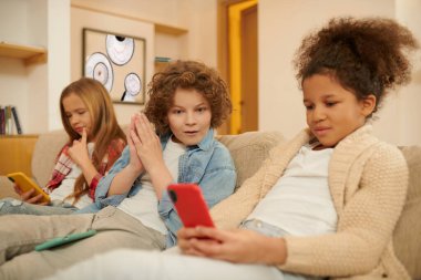On internet. Kids sitting on the sofa with gadgets in hands and looking involved