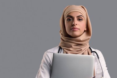 Kadın bir doktor. Başörtüsü ve laboratuvar önlüğü giymiş dizüstü bilgisayarlı bir kadın.