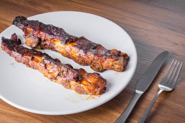 Beyaz bir porselen tabakta barbekü soslu kızarmış domuz kaburgası, grafit ve bıçakla. Tahta masa. Perspektif görünümü.
