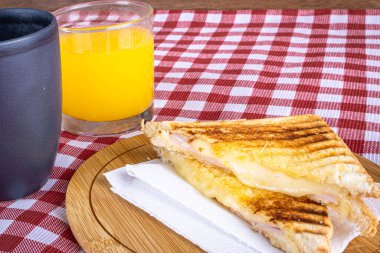 Grilled ham and cheese. Sandwich with cheese and ham on the grill. Served on a checkered tablecloth. Accompanied by orange juice. Perspective view.