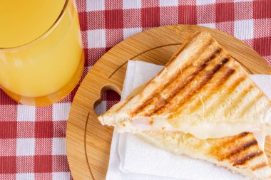 Grilled ham and cheese. Sandwich with cheese and ham on the grill. Served on a checkered tablecloth. Accompanied by orange juice.