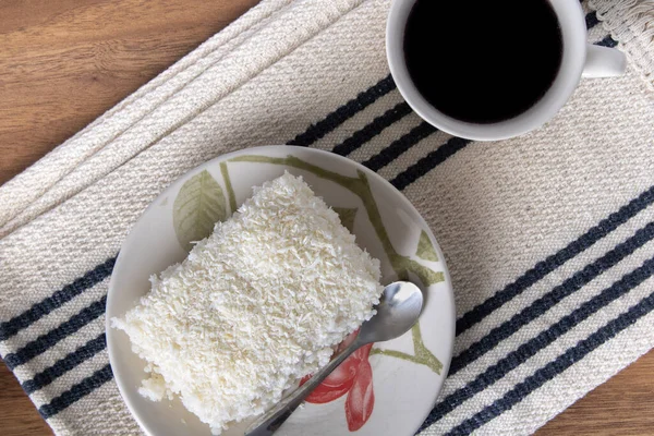 Piece of couscous in porcelain saucer on wooden table, typical Brazilian dish, made with tapioca, milk, sugar and coconut milk. With coffee cup. Perspective view.