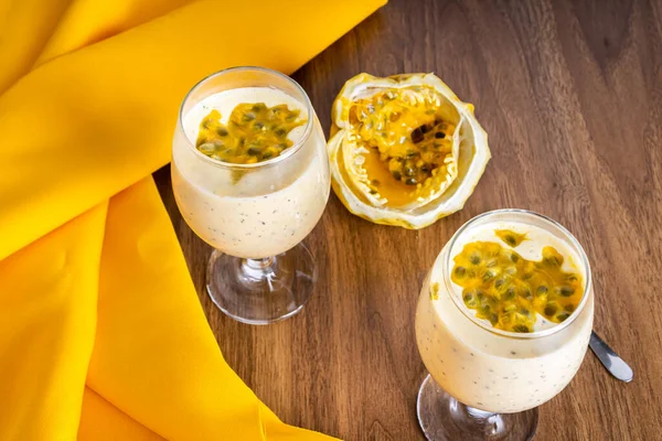 Passion fruit mousse in a glass bowl, with passion fruit in the background, on a wooden table and a yellow tablecloth around it. Perspective view.