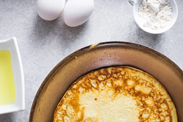 stock image Delicious pancakes in a frying pan on a white table, with wheat, eggs, olive oil, tomato sauce and a checkered tablecloth in the background. Top view.