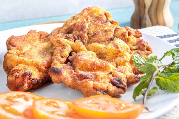 stock image Delicious Milanese chicken (breaded chicken with breadcrumbs and egg), a typical Brazilian dish. On a white plate with tomatoes, lemon on striped white tablecloth and blue marble. Close view.