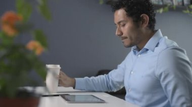 Kahverengi tenli ve kıvırcık saçlı genç bir adam iş yerinde tabletini ve kahvesini evde kullanıyor.